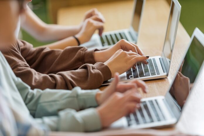 students-typing-on-laptops-GettyImages_1513744777.jpeg High school students sitting at the desk in the classroom during lesson, using laptops. Close up of hands, unrecognizable people.