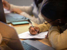 A student focuses on completing a worksheet with a pen during class, with a laptop open in front of them.