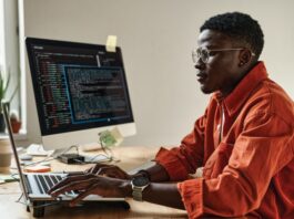 A man sits at a computer desk looking at a monitor. He’s wearing glasses, and the sleeves of his orange shirt are rolled up.
