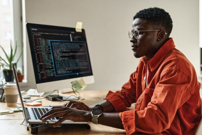 A man sits at a computer desk looking at a monitor. He’s wearing glasses, and the sleeves of his orange shirt are rolled up.