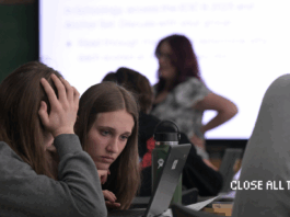 Two female high school students hover over a computer. One student has a hand covering her face as the other student leans in closely to the computer screen.Their teacher and a projection of the lesson are blurred in the background. The Close All Tabs logo appears in pixelated font in the lower right corner.