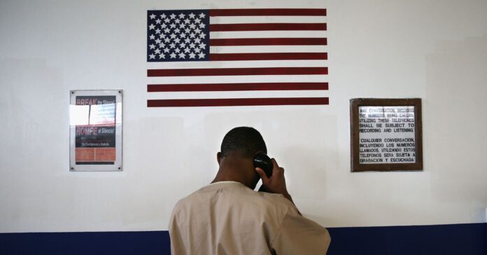 YWwuanBn.jpeg A Black man in a tan prison uniform stands in front of a black and white wall, holding a phone to his ear. A U.S. flag and two signs are on the wall. One sign reads, "Break the silence!" in English and Spanish. The second sign details that calls will subject to recording and listening.