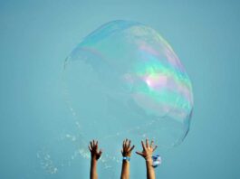 hands of children reach up to giant soap bubble as it pops against blue sky
