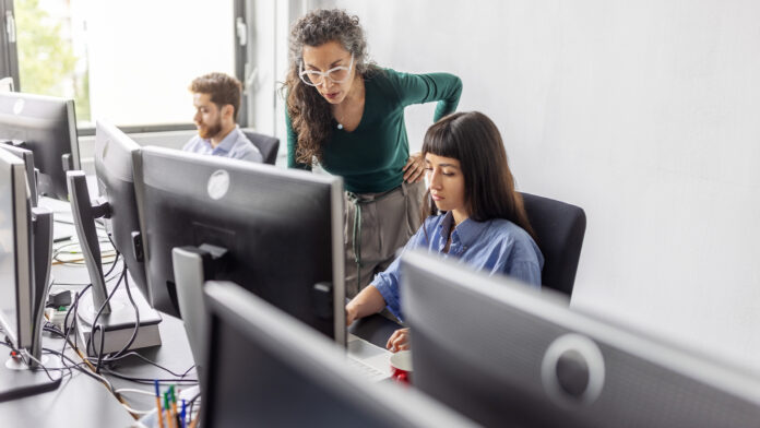 AHK2hBouv4dyfiwnu25mQo.jpg Male and female software developers working on desktop computers in an open plan office space with senior employee leaning over and observing work.