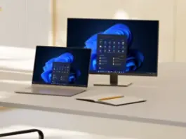 An open laptop and monitor sit on a desk alongside a notebook and pencil, with both screens displaying Windows bloom backgrounds and open start menus