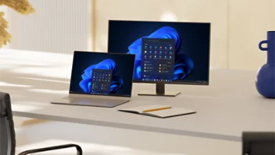 An open laptop and monitor sit on a desk alongside a notebook and pencil, with both screens displaying Windows bloom backgrounds and open start menus