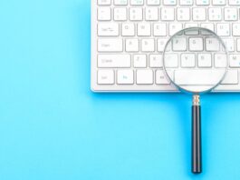 a photo of a keyboard on a blue background, with a magnifying glass on top of the keyboard (gettyimages-1372176504)