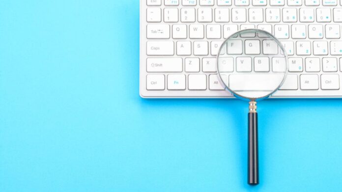 gettyimages-1372176504.jpg a photo of a keyboard on a blue background, with a magnifying glass on top of the keyboard (gettyimages-1372176504)
