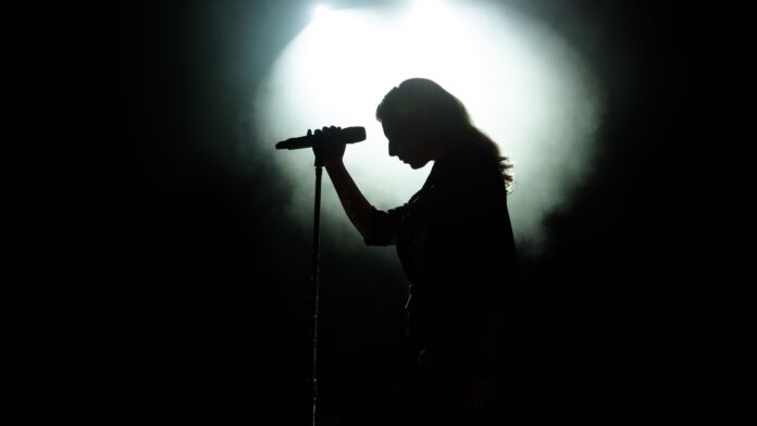 hb89B7aMthcdUR3mfh89FX.jpg a dramatically-lit photograph showing the silhouette of a woman with a microphone