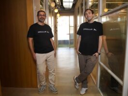 The two co-founders stand abreast in an office hallway wearing black logo t-shirts.