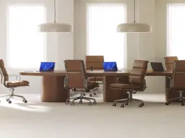 Three open laptops, with two displaying Windows blue bloom backgrounds, sit on a long wood conference room table surrounded by six brown leather office chairs
