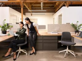 Photograph of two female executives in an office. One executive is seated, holding a Surface Laptop 13" Platinum device in her right hand. A Surface Pro 12" Platinum device in laptop mode is on another desk, connected to an external monitor. A Surface Arc Mouse is next to the device.