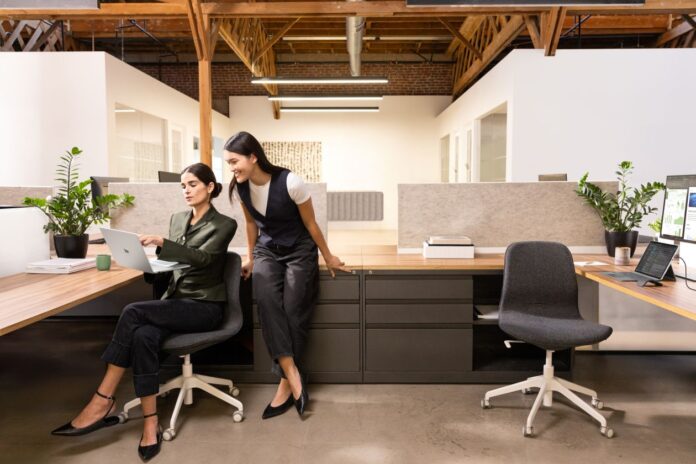 SUR25-COMMR-LaptopPro-Snapdragon-Platinum-Office-Collab-01.jpg Photograph of two female executives in an office. One executive is seated, holding a Surface Laptop 13" Platinum device in her right hand. A Surface Pro 12" Platinum device in laptop mode is on another desk, connected to an external monitor. A Surface Arc Mouse is next to the device.