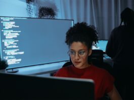 Female software developer coding on a desktop computer in a dimly-lit office space with computer code pictured on large screen in background.