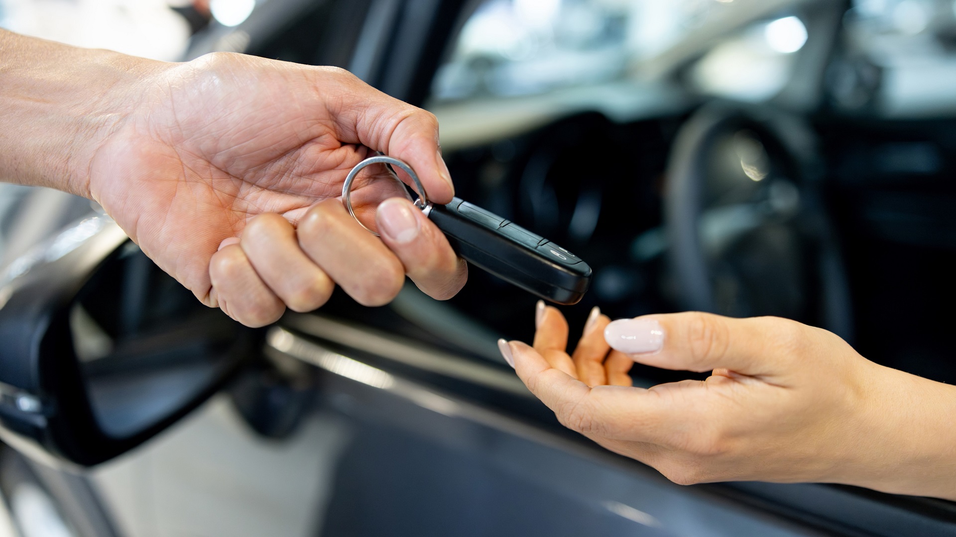 handing-vehicle-keys-sell-car-concept_iStock-1977609685.jpg Woman sitting in a car and reaching her hand out to receive the keys from the salesperson at the dealership
