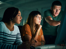 Man standing over two younger people and pointing to the computer screen they are all looking at. 