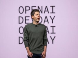 An individual is standing on stage with "OPENAI DEMO DAY" in the background, wearing a dark green sweater and dark pants, presenting to an audience.