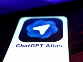 A smartphone screen displaying the ChatGPT Atlas app icon, which features a blue, swirling design with a white paper airplane symbol in the center. The phone is held against a dark, out-of-focus background with hints of purple light.