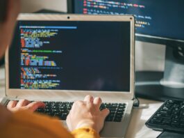 Software developer using AI coding tools on a laptop computer with source code pictured on screen and desktop monitor in background.