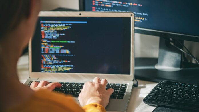 pXaDHxpCwz8PrGomizBmrT-1920-80.jpg Software developer using AI coding tools on a laptop computer with source code pictured on screen and desktop monitor in background.