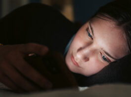 A teenage girl lies on her side in the dark, her face illuminated by her smartphone, suggesting she is interacting with something on the screen.