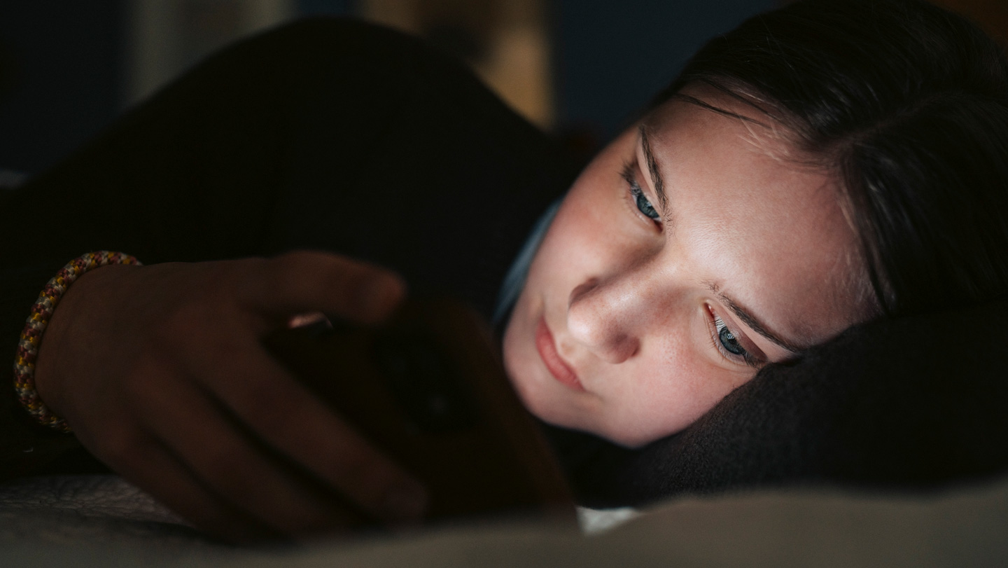 102925_LS_chatbots_feat.jpg A teenage girl lies on her side in the dark, her face illuminated by her smartphone, suggesting she is interacting with something on the screen.
