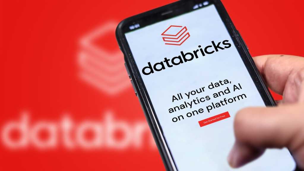 4085696-0-86482200-1762428532-databricksphone.jpg A photograph of a Databricks website on a phone being held in front of a larger, blurry version of the Databricks logo.