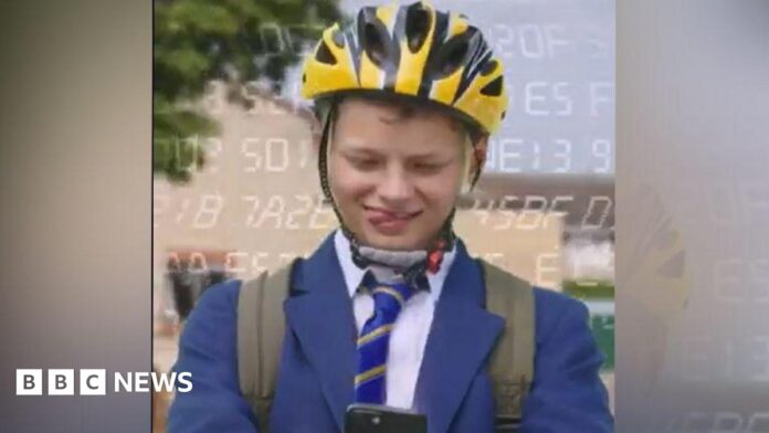 A boy in a blue blazer and striped tie has a yellow helmet on and a rucksack, and he is looking down at a phone. He is sticking his tongue out to one side of his mouth. He is outside.