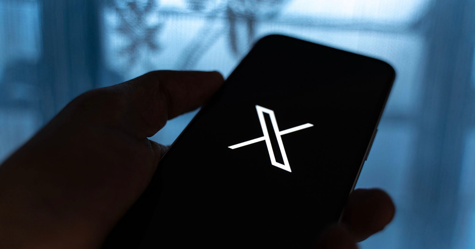 X-Twitter-Elon-Musk.jpg A hand holds a smartphone displaying a white "X" logo on a black screen, with a blurred blue background and some silhouetted shapes visible.