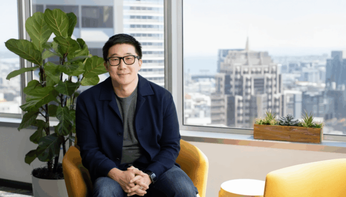 A man in glasses and a dark blue jacket sits in a yellow chair in an office with a city view.