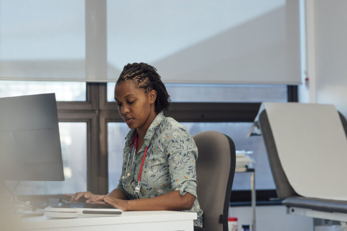Side view shot of a female general practitioner sitting at her desk with a computer, typing on her keyboard.
