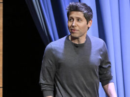 A man in a gray sweater stands on a stage with blue curtains, looking to the side.