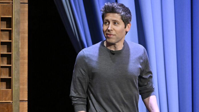 A man in a gray sweater stands on a stage with blue curtains, looking to the side.