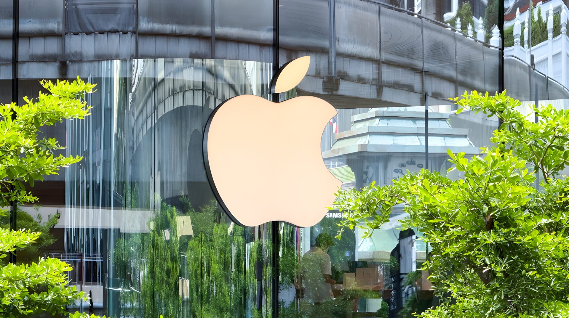 Apple-1-1920x1074.jpg A large Apple logo is displayed on the glass facade of an Apple Store surrounded by greenery and reflecting nearby buildings.