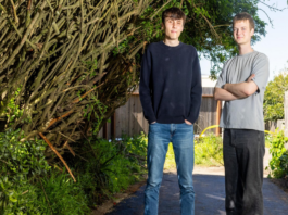 Two young men stand on a paved path between dense green bushes and a wooden fence, with one wearing a black sweater and jeans and the other in a gray shirt and black pants.