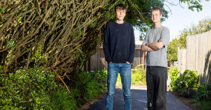 Two young men stand on a paved path between dense green bushes and a wooden fence, with one wearing a black sweater and jeans and the other in a gray shirt and black pants.
