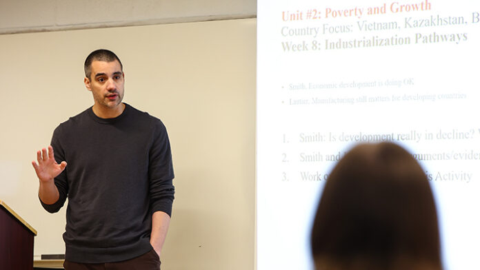 Professor teaching in a classroom in front of slide projected on screen.
