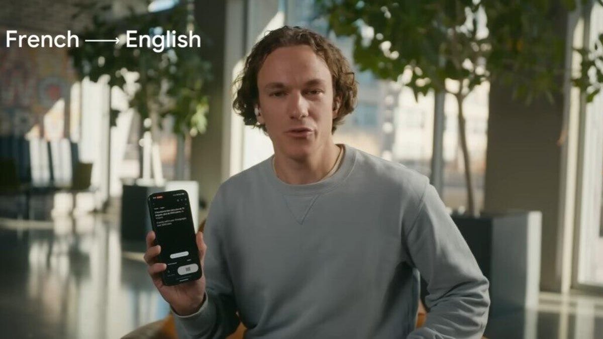 google-headphonetranslate.jpg A man in a gray sweater holds up a phone that's doing live translation of French to English language. He's wearing earbuds.