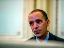 “OpenAI Policy Chief Warns ‘Doomers’ Are Treading Dangerous Ground” – The San Francisco Standard A man with short dark hair and a serious expression wears a dark suit and light blue shirt, looking slightly downward in a softly lit room.