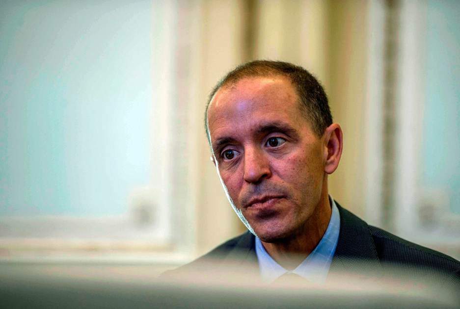 1776263440_S1200x630-FPNG.png A man with short dark hair and a serious expression wears a dark suit and light blue shirt, looking slightly downward in a softly lit room.