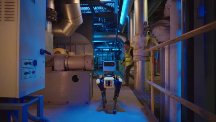 A four-legged Boston Dynamics robot called Spot walks down the hallway of an industrial facility. A human worker wearing a bright yellow safety vest is starting to cross the hallway behind the robot.